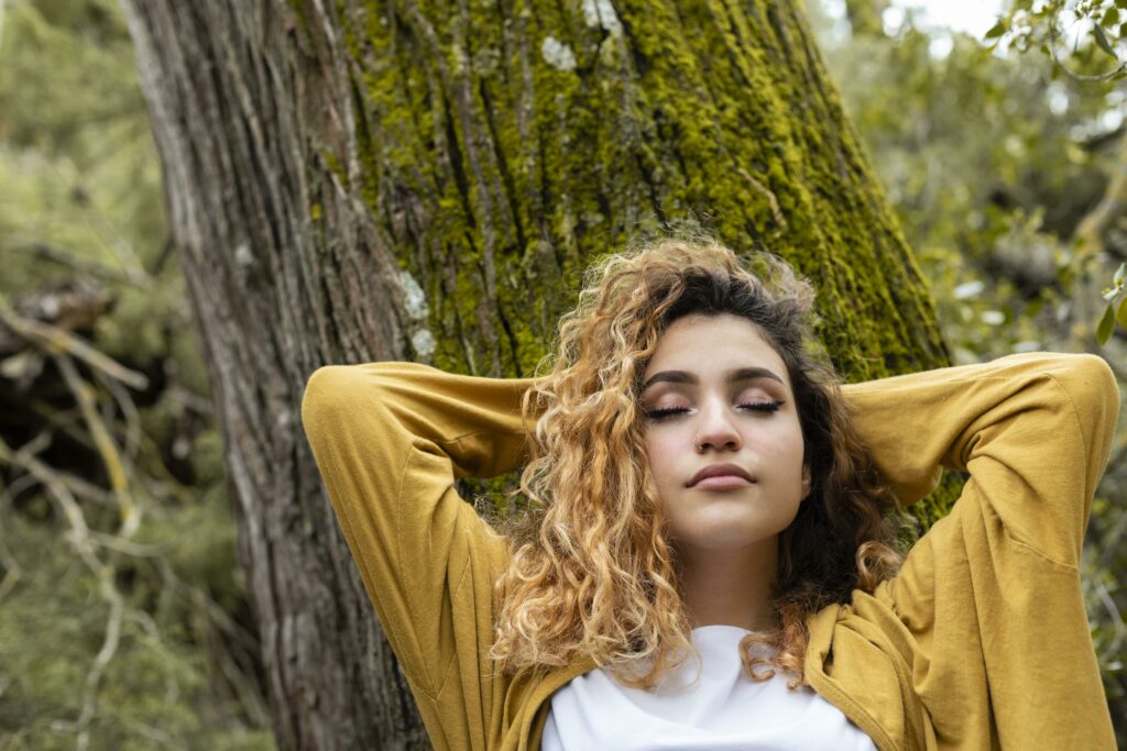 Sophrologie, une femme est assise contre un arbre avec les yeux fermés et les mains croisées derrière la tête, inspirant le bien-être et les bienfaits de l'activation du nerf vague