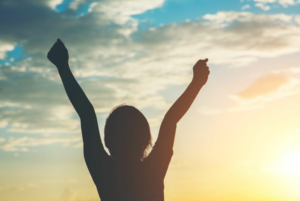 silhouette d'une femme levant les bras en signe de reussite et de gratitude face au coucher de soleil